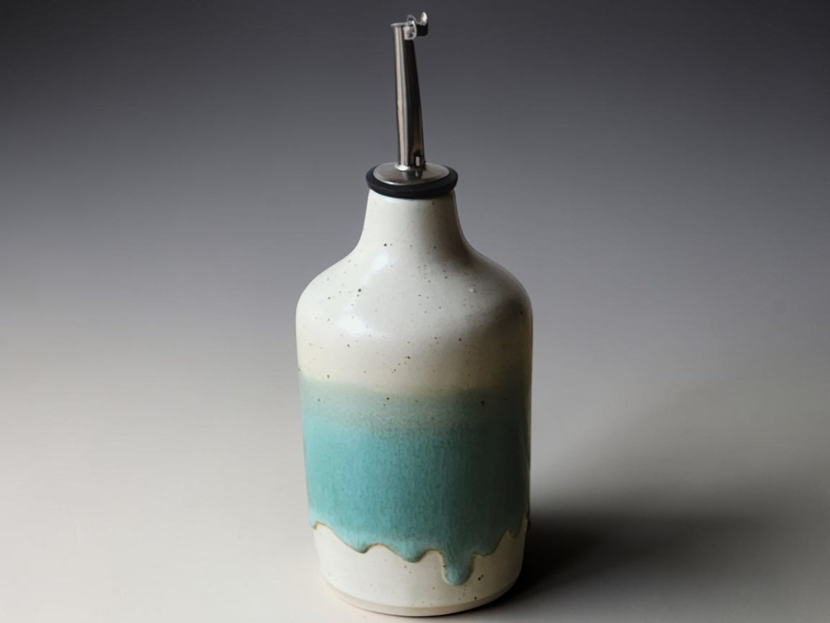 A ceramic oil dispenser bottle with a metallic pour spout, featuring a speckled off-white glaze that fades into a turquoise blue gradient at the bottom with a dripping effect.