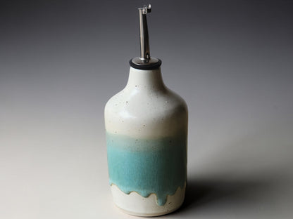 A ceramic oil dispenser bottle with a metallic pour spout, featuring a speckled off-white glaze that fades into a turquoise blue gradient at the bottom with a dripping effect.