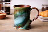 Handmade ceramic mug with green teal drip glaze and rust interior on timber surface