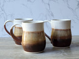 Three handmade ceramic mugs with earthy brown and cream glaze, wheel-thrown stoneware pottery