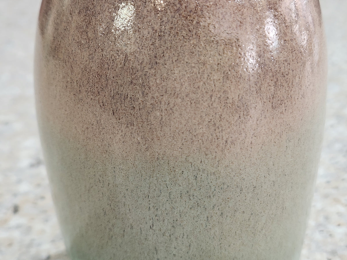 Close-up of a ceramic vase with a speckled glaze that transitions from a warm taupe-brown at the top to a muted sage green at the bottom, set against a light speckled stone surface.