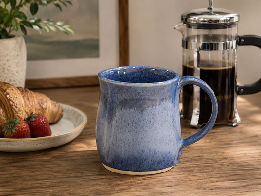 Handmade blue pottery mug with French press coffee and croissant on timber table