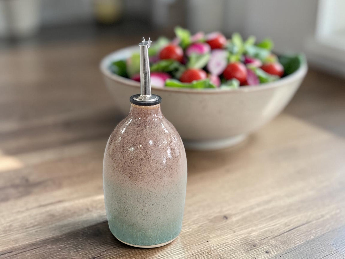 A ceramic oil dispenser bottle with a pink and green gradient glaze, featuring a clear glass pour spout, set on a wooden table with a blurred salad bowl in the background.