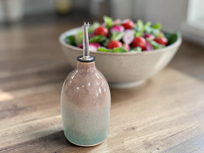 A ceramic oil dispenser bottle with a pink and green gradient glaze, featuring a clear glass pour spout, set on a wooden table with a blurred salad bowl in the background.