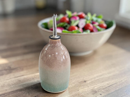 A ceramic oil dispenser bottle with a pink and green gradient glaze, featuring a clear glass pour spout, set on a wooden table with a blurred salad bowl in the background.
