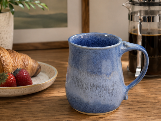 Blue ceramic mug on a wooden table with a croissant, strawberries, and a French press in the background.