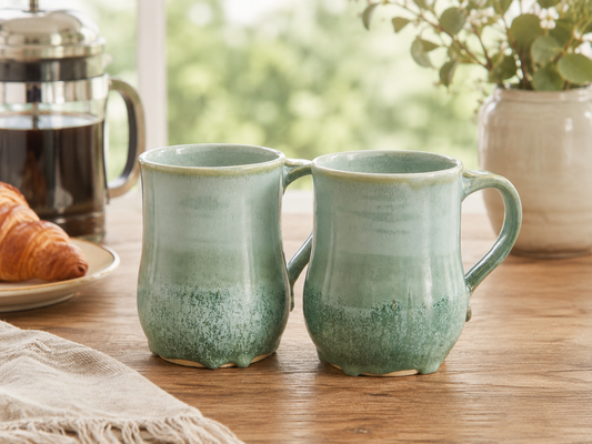 Pair of large green stoneware mugs beside a French press and croissant