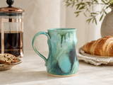 Handmade ceramic mug in Bush Creek glaze styled with French coffee press and pastries in a warm kitchen setting
