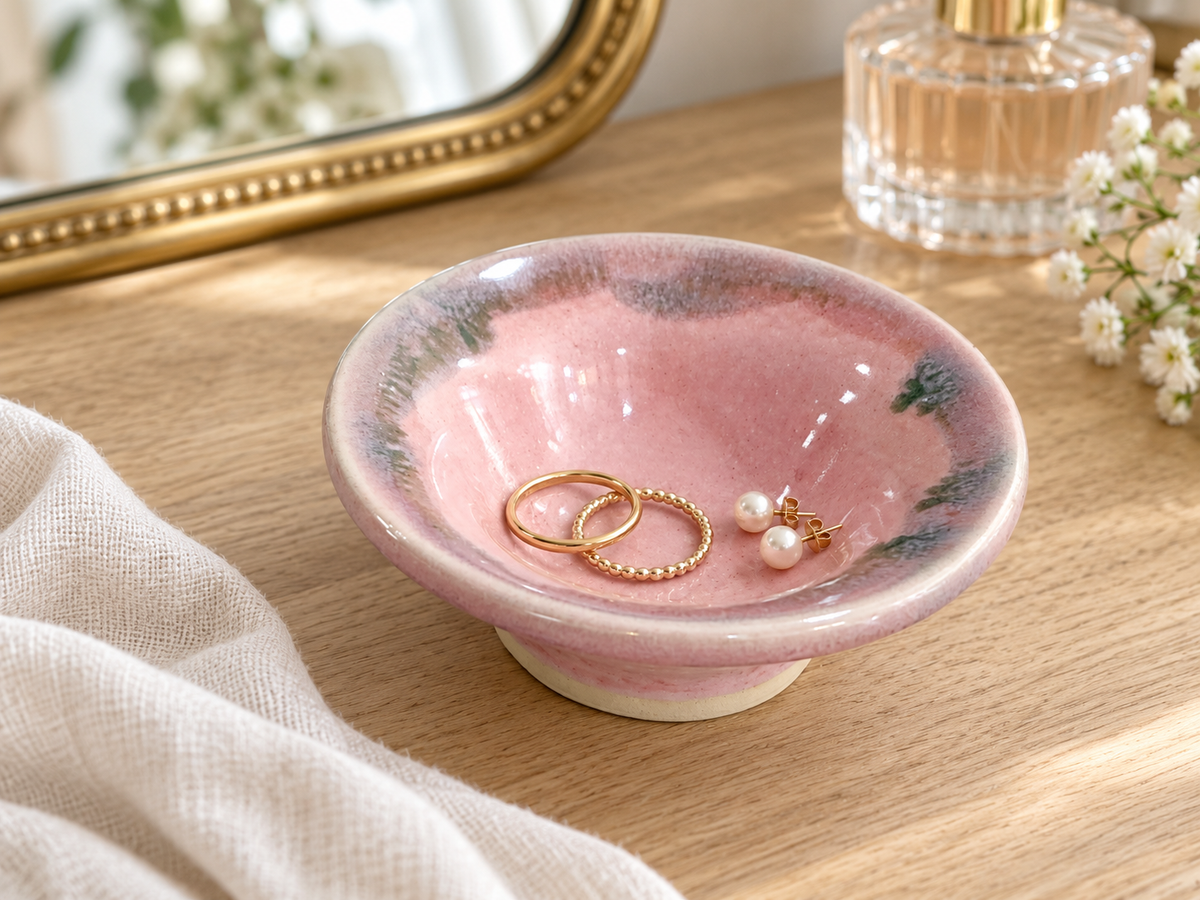 Pink pottery jewellery bowl holding gold rings and pearl earrings on a timber dressing table.