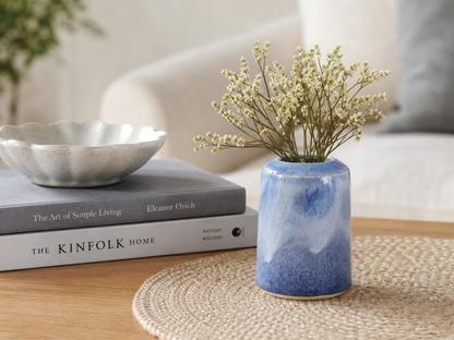 Handmade blue ceramic bud vase with small white flowers styled on a woven mat with books in a neutral home setting