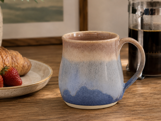 Handmade pottery mug with oatmeal and blue mottled glaze styled beside French press coffee and breakfast pastry