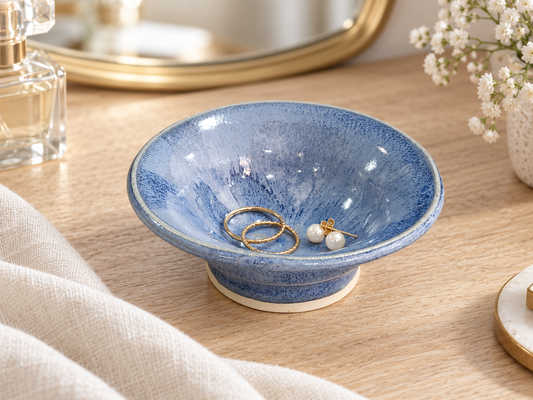 Blue pottery jewellery bowl holding gold rings and pearl earrings on a timber dressing table.