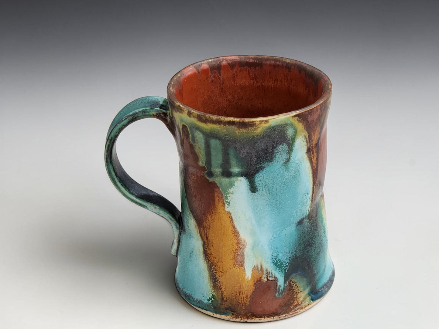 Opposite side view of a handmade ceramic mug with layered bush-inspired glazes in earthy red, aqua and moss green.