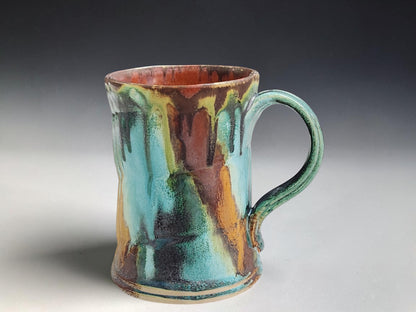Handmade pottery mug with terracotta interior and layered turquoise, green and earthy brown glaze, front view on a neutral background.