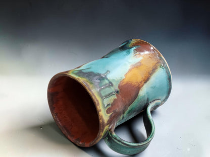 Angled studio view of a handmade ceramic mug with curved handle and rich terracotta, turquoise and green glazes.