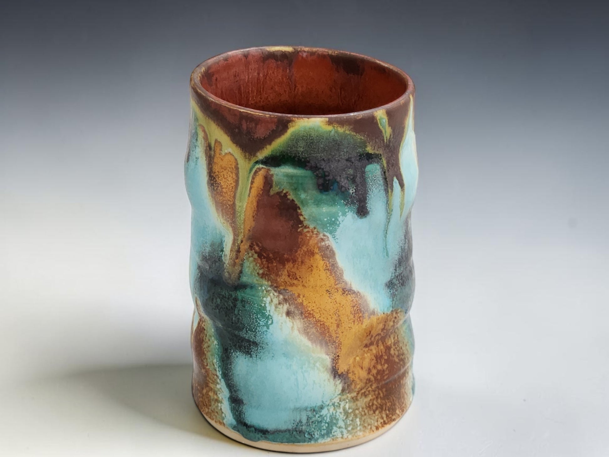 Handmade ceramic coffee mug in turquoise, rust brown and moss green glaze, photographed on a neutral grey studio sweep