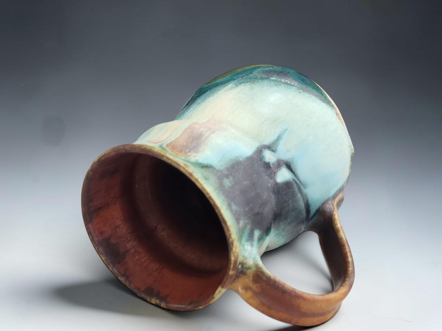 A handmade ceramic mug with a handle, featuring a marbled glaze of light blue, teal, brown, and dark charcoal, shown on a gray gradient background.
