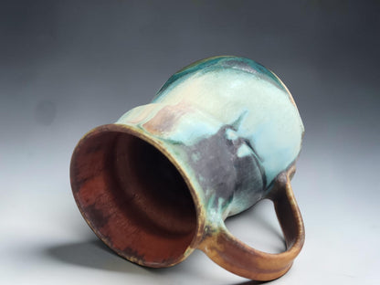 A handmade ceramic mug with a handle, featuring a marbled glaze of light blue, teal, brown, and dark charcoal, shown on a gray gradient background.