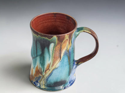 Tall handmade coffee mug with flowing blue-green glaze and cream-edged ochre patterns.