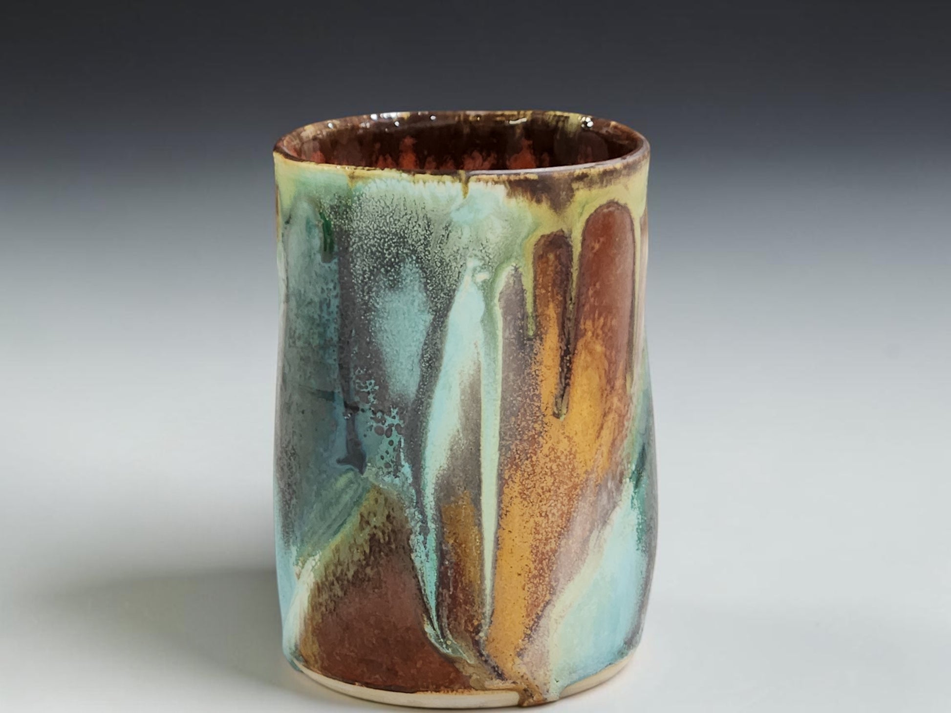 A handmade ceramic cup with a marbled glaze featuring brown, teal, and ochre orange colors, set against a neutral gray background.