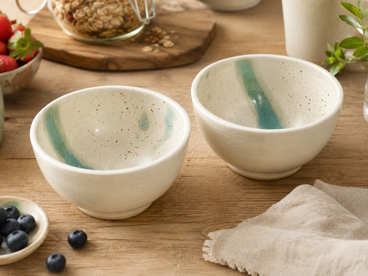 Handmade ceramic cereal and soup bowls in speckled cream glaze with green accent, styled with breakfast berries in a warm kitchen setting