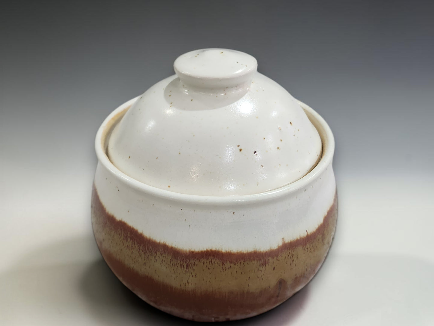 Handmade ceramic sugar bowl with lid featuring cream and warm brown glaze, photographed on a professional studio sweep with a neutral grey gradient background.