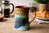 Handmade pottery mug in galaxy glaze with French press and croissant on kitchen table