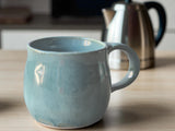 Blue ceramic mug on a wooden surface with a coffee pot in the  background