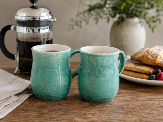 Handmade green crystalline porcelain mug with coffee and croissant on a timber table