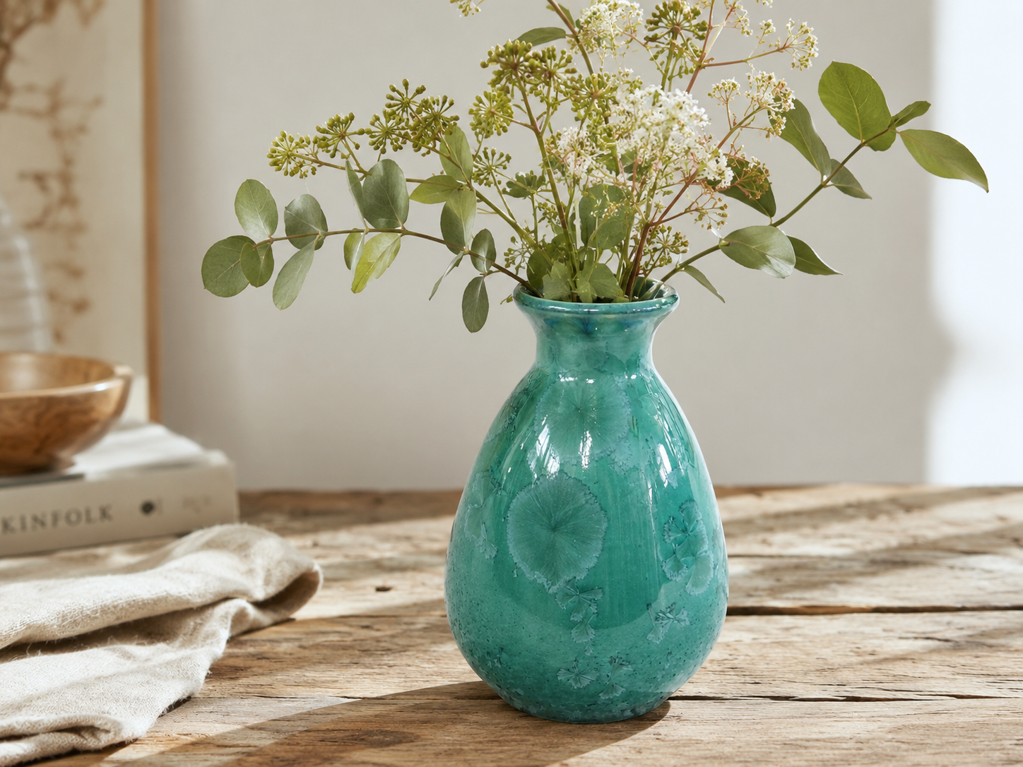 Green crystalline porcelain vase with foliage and white flowers on a timber table