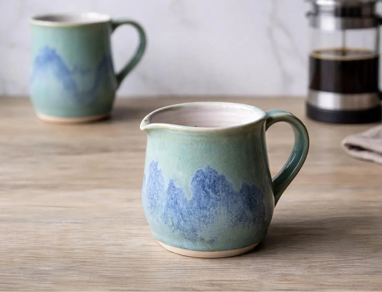 A ceramic creamer pitcher with a blue mountain landscape glaze sits on a wooden table, with a matching mug in the background and a French press blurred behind it.