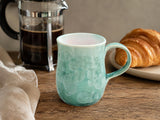 Handmade crystalline glazed green ceramic mug on timber table with French press coffee and croissant
