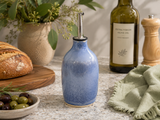 Handmade blue ceramic olive oil bottle with pourer on kitchen bench beside artisan bread and olives