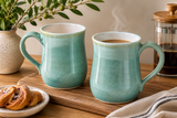 Handmade ceramic mugs by Buxton Pottery featuring a mottled green glaze and glossy white interior, styled in a warm coffee setting and handcrafted in Melbourne, Australia.