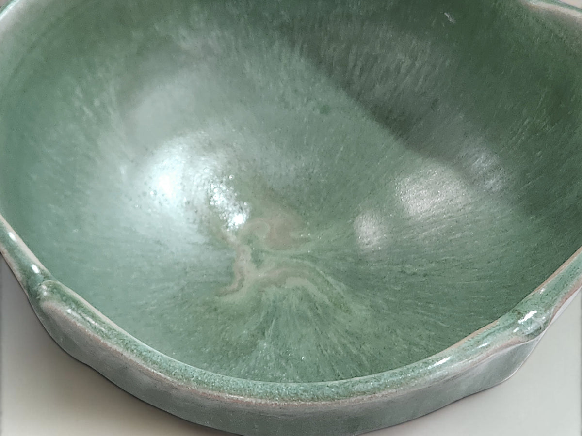 A close-up of a ceramic bowl with a glossy, swirled sage green glaze and a flared rim, set against a plain light background.