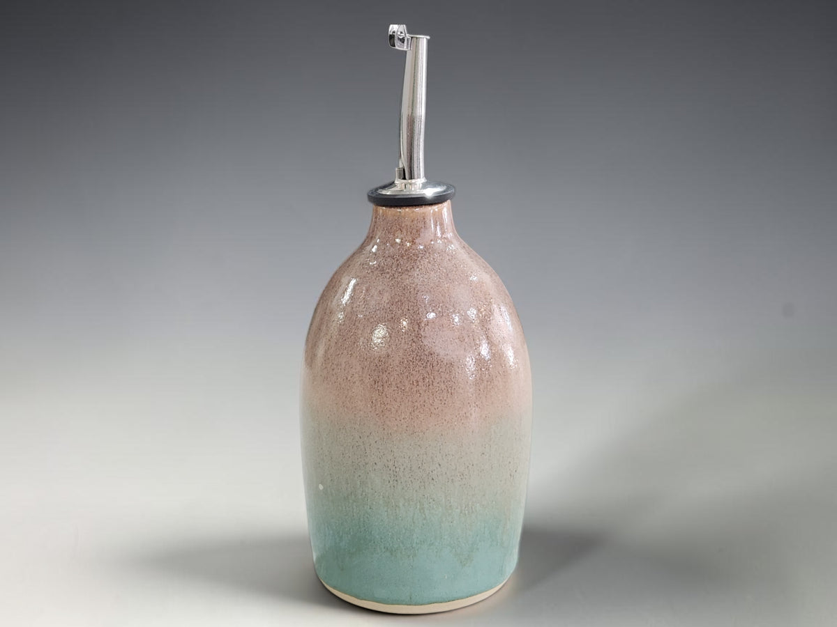 A ceramic oil dispenser bottle with a glossy gradient glaze transitioning from soft brown at the top to light green at the bottom, featuring a silver metal pour spout.