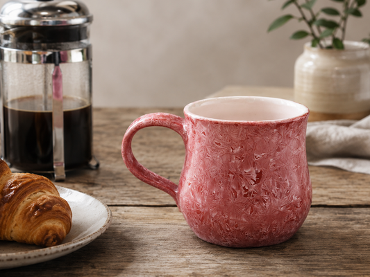 Red crystalline porcelain mug with French press and croissant on a timber table