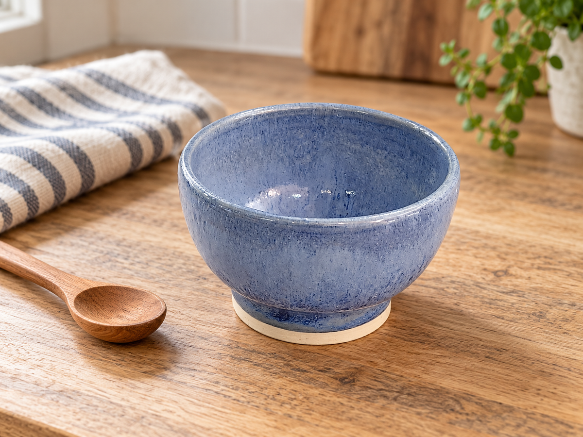 Small blue pottery bowl on a timber benchtop beside a wooden spoon and striped linen.