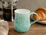 Handmade crystalline glazed green ceramic mug on timber table with French press coffee and croissant