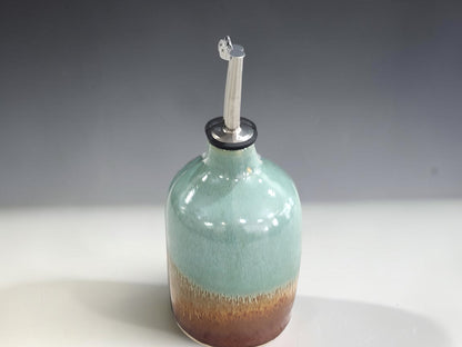 A ceramic oil bottle with a glossy light blue-green glaze that fades to a warm brown at the base, featuring a silver metal spout , set against a neutral gray background.