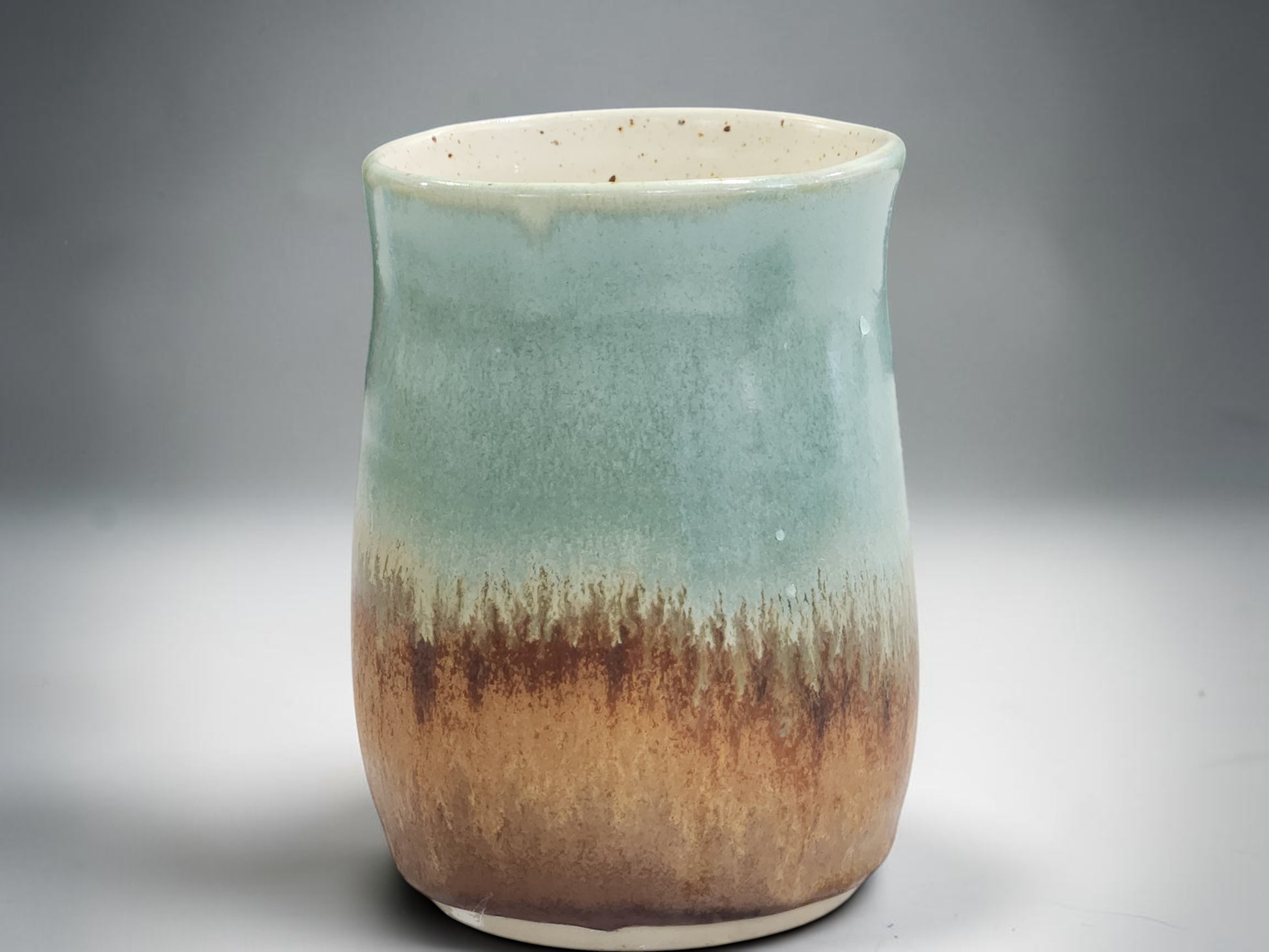 A handmade ceramic cup with a glossy glaze featuring a gradient transition from light teal-green at the top to a rustic brown at the bottom, set against a neutral gray background.